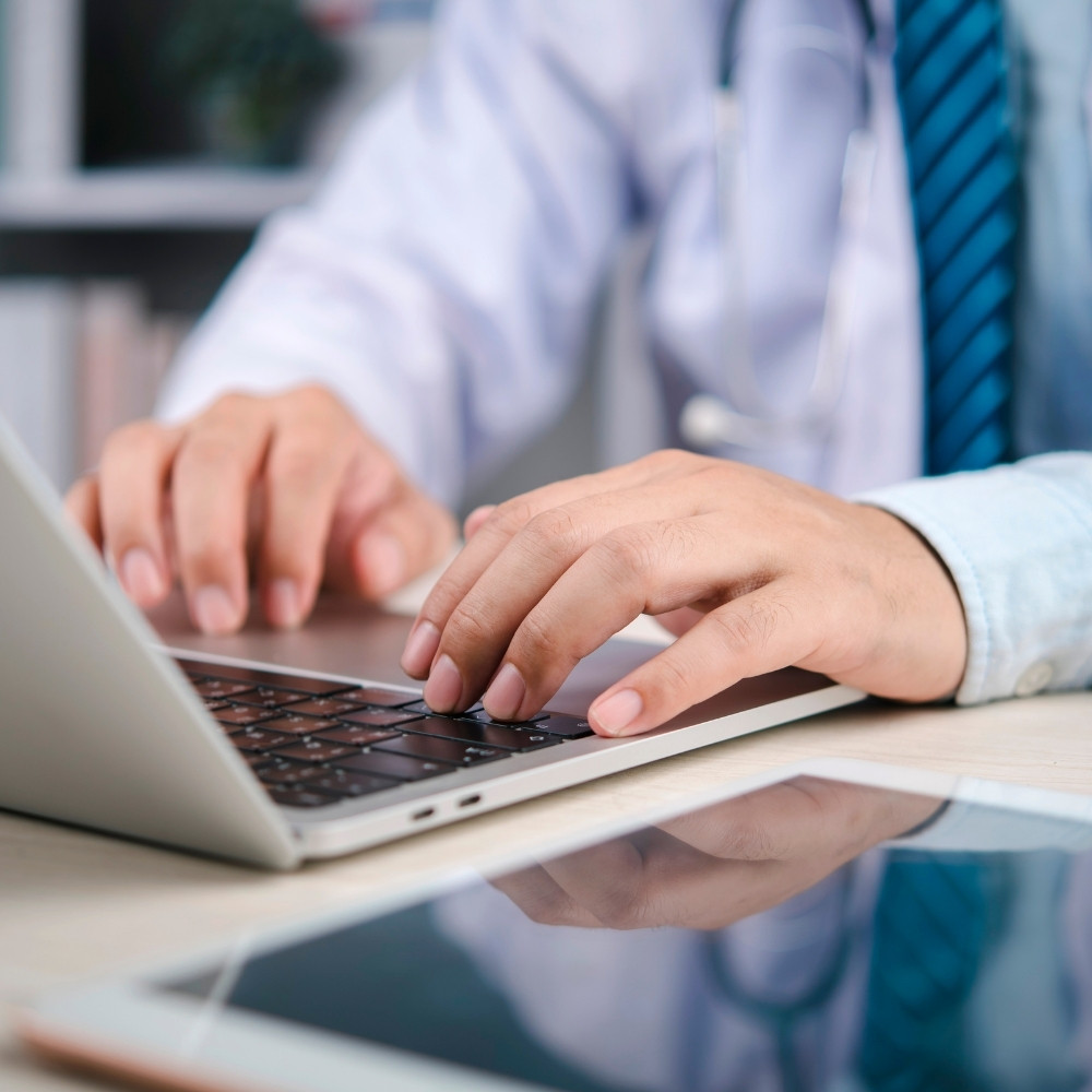 ein arzt sitzt an seinem laptop und tippt etwas auf der tastatur. er trägt ein stetoskop. auf dem tisch liegt noch ein tablet. zum thema studie zur elektronischen patientenakte ePA